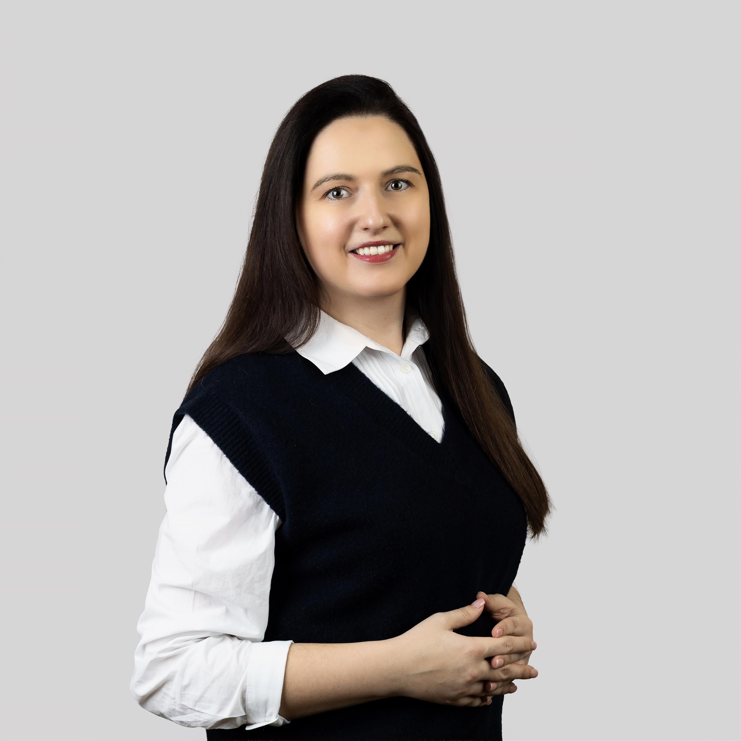 Indrė Baltrušaitienė | KTU Centre for Teacher Education “iSTEAM” Portrait of a smiling woman with long dark hair, wearing a white shirt and a black sleeveless top, against a light grey background.
