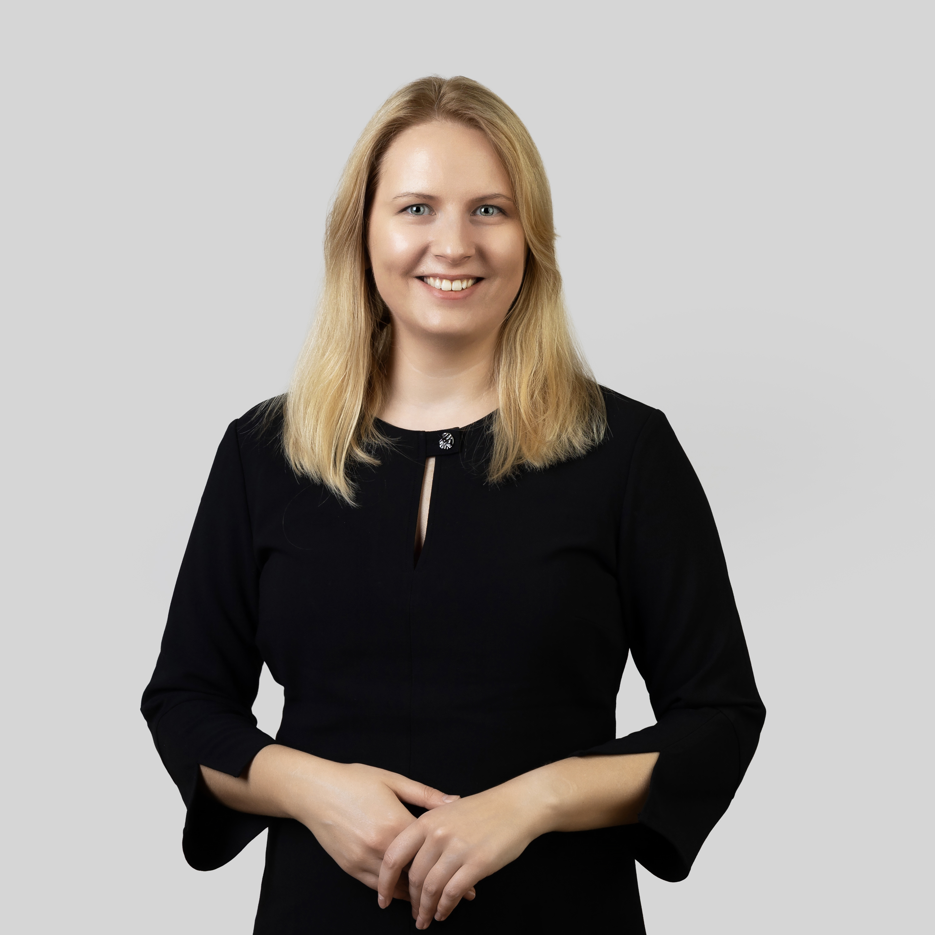 Akvilė Grigaliūnė | KTU Centre for Teacher Education “iSTEAM” Portrait of a smiling woman with shoulder-length blonde hair, wearing a black dress and standing with her hands clasped against a light grey background.