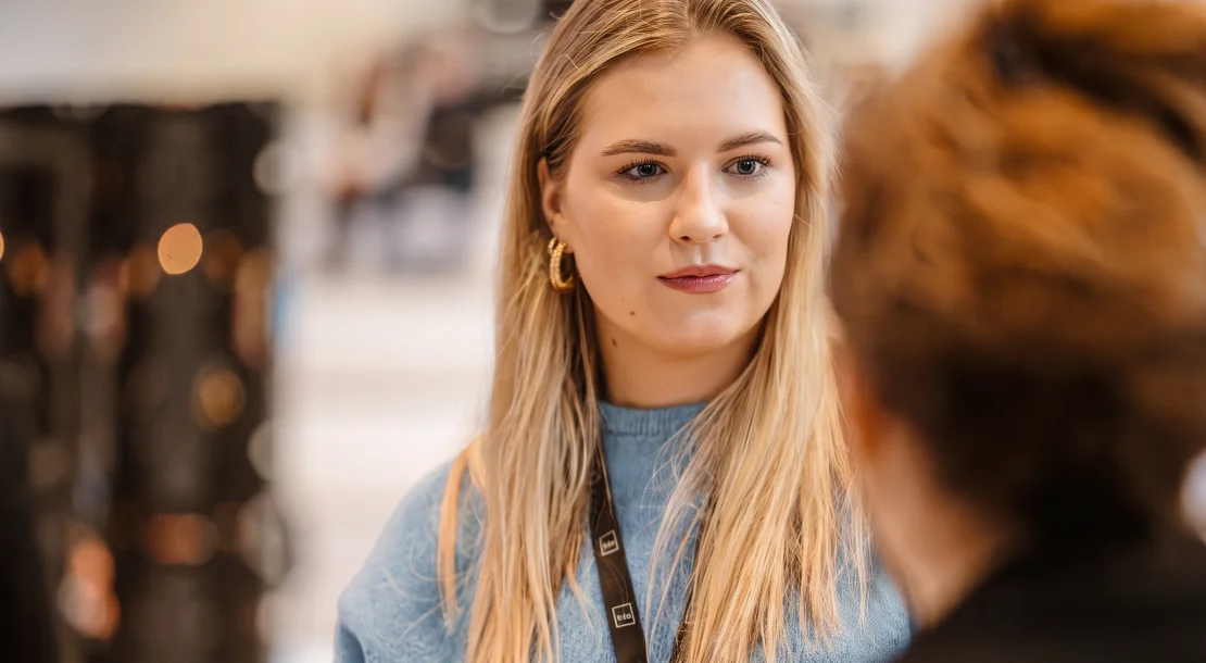 Young woman with long blonde hair wearing a blue jumper, engaged in conversation at an indoor event.