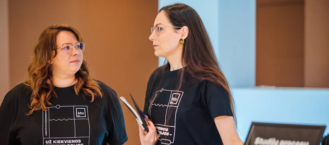 Consultations Two women wearing KTU T-shirts having a conversation at a university event, one holding documents.