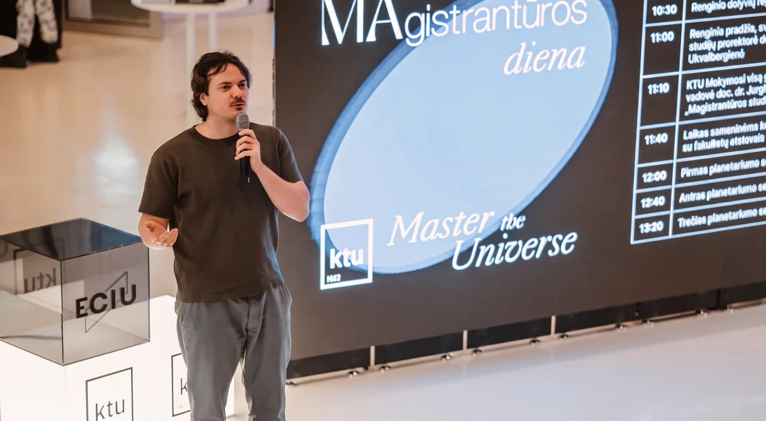 Speaker holding a microphone during KTU Master's Day, standing in front of a presentation screen displaying the event programme.